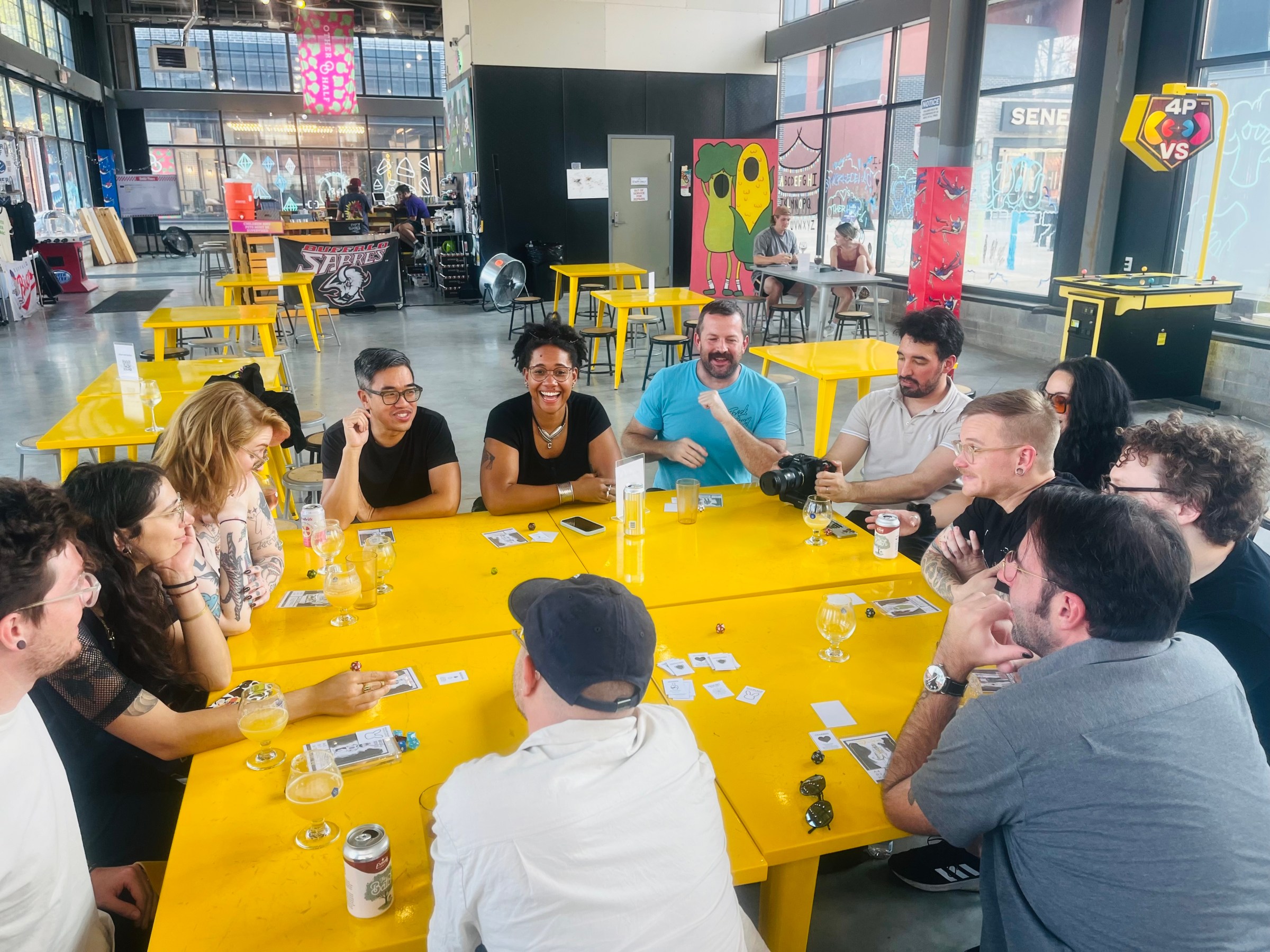 The creative team behind the delicate comic series, sitting around a large yellow table playing a prototype of Corpse: The Game.